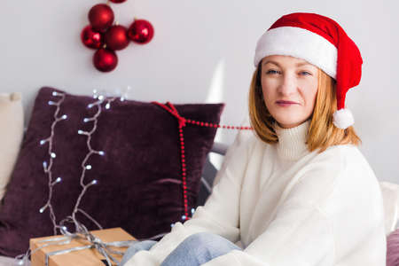 A young woman in a white sweater, Santa Claus hat, with New Year gifts sits on the sofa, looks at the camera and smilesの写真素材