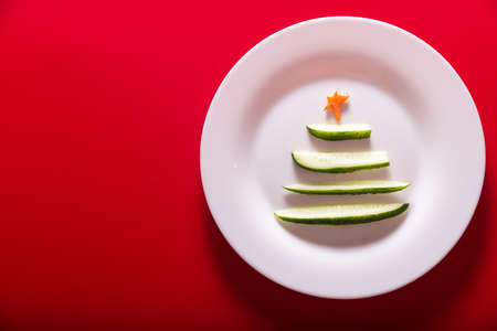 New Year cucumber in the form of a Christmas tree on white plate on red background. Top view. Copy space. Horizontal photoの写真素材