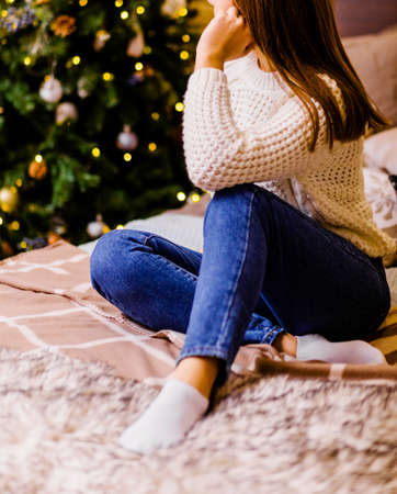 Young woman in blue jeans and white knitted sweater sits on the bed and looks at the christmas tree. Photoの写真素材