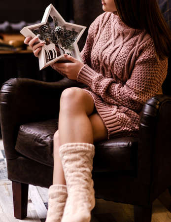 A young woman in a pink knitted sweater and socks sits in an armchair and holds a wooden star-shaped decor in her hands. Photoの写真素材
