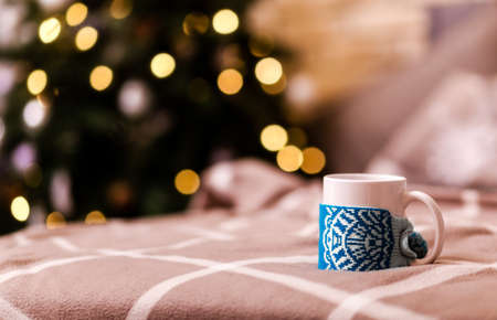 A white cup in winter decor stands on a brown plaid plaid against the background of a Christmas tree with a garland. Photoの写真素材