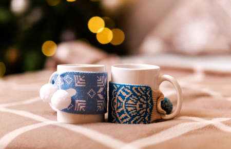 Two white cups in winter decor stand on a brown plaid plaid against the background of a Christmas tree with a garland. Romantic new year concept. Photoの写真素材