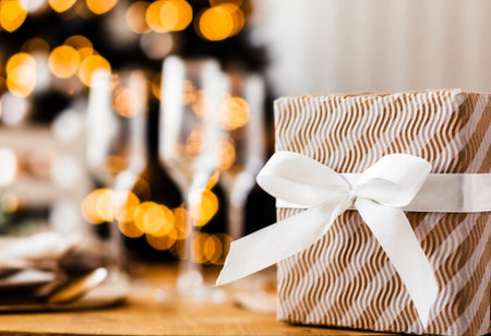 A gift box with a bow stands against the background of a Christmas tree and glasses of champagne....