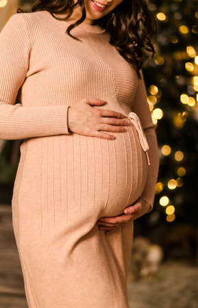 The hands of a pregnant woman lie on her stomach. Woman posing against the background of a christmas tree. Photoの写真素材