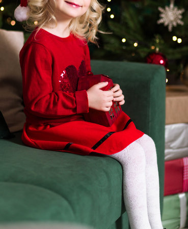A little girl in a red dress and white tights sits on a green sofa with a toy house in her hands....