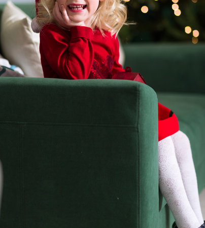 Little girl in a red dress sits on a green sofa, smiles and looks at the camera. Photo
