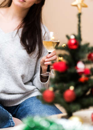 A beautiful brunette woman is sitting in the room on the couch next to a Christmas tree decorated with toys. A girl holding a glass of wine in her handの写真素材