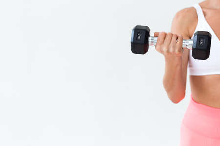 A brunette girl in a white top and in coral leggings makes lunges with her legs with dumbbells in her hands against a white wall. Vertical photoの写真素材