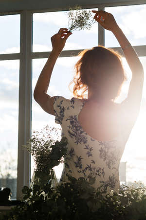 The girl is sitting in front of the window with a bouquet of flowers in her raised hands. Outside the window is a warm summer eveningの写真素材