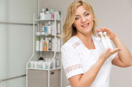 A woman doctor cosmetologist holds a dispenser with a cosmetic product in her hands, looks into the camera and smiles. Cosmetologist posing in a beauty parlorの写真素材