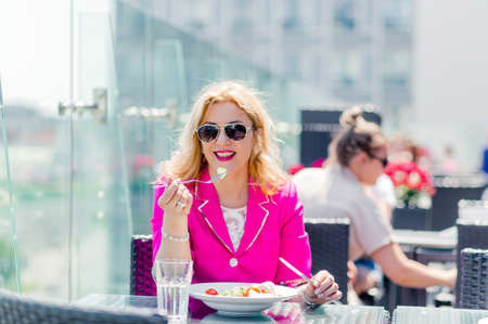 Beautiful woman in a bright magenta jacket sits on a summer terrace and eats a fresh vegetable salad. Horizontal photoの写真素材