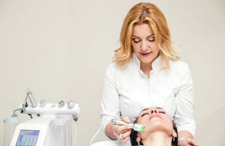 The beautician makes the patient the procedure of microcurrent therapy. Next to the beautician is a...