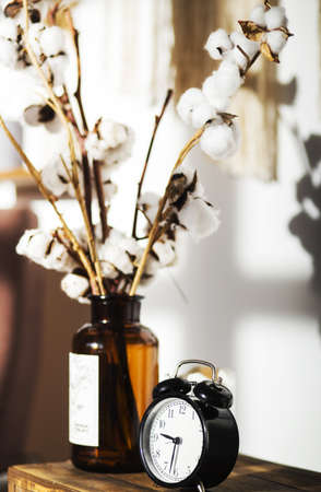 There is a black retro clock and a vase with cotton branches on a wooden shelf. Vertical photoの写真素材