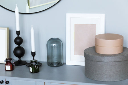 There is a chest of drawers near the gray wall with many interior details. There is a mirror on the wall, candles, boxes and a photo frame on the dresserの写真素材