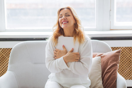 A woman in white clothes reads on the couch at home and hugs a piece of paper with her wishes....