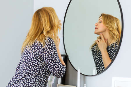 A beautiful mature woman in a fashionable dress stands in front of a mirror, touches her skin and assesses its condition. Woman happy life concept after 40 yearsの写真素材