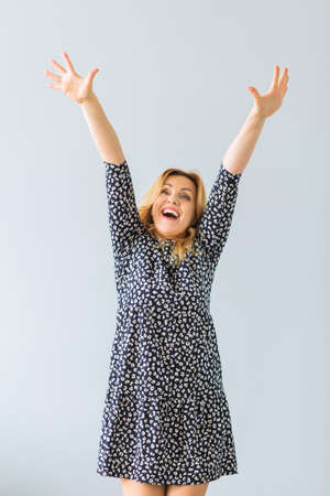 A beautiful mature woman in a fashionable dress stands in front of a gray wall with raised hands. Woman happy life concept after 40 yearsの写真素材
