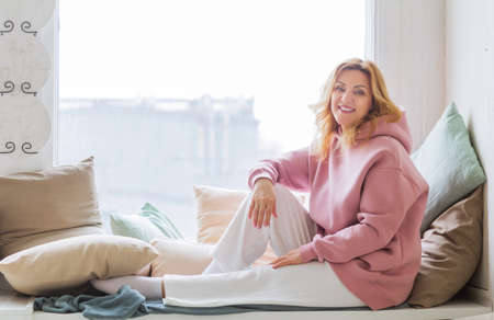 A mature woman in a pink hoodie sits on the windowsill, looks at the camera and smiles. Woman happy life concept after 40 yearsの写真素材