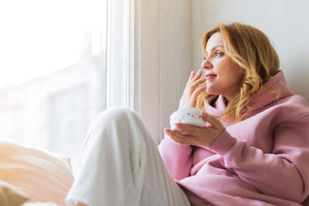 A mature woman in a pink hoodie sits on the windowsill, holds a jar of cream in her hands and sniffs it. The concept of a beautiful and healthy lifeの写真素材