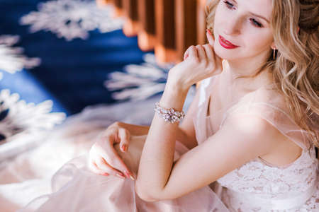 A beautiful stylish bride in a long light pink wedding dress poses in a bright hotel lobby sitting on a carpet near a wooden wall. Bride portraitの写真素材