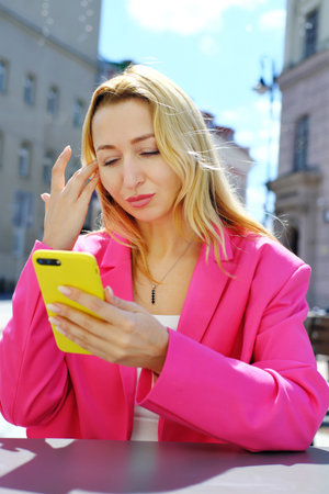 A beautiful adult blonde woman in a bright fuchsia-colored jacket sits at a table in a cafe and...