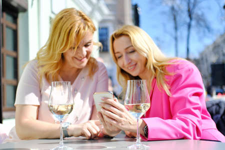 Two beautiful fashionable adult woman girlfriends are relaxing in a cafe in the city center on a summer terrace, drinking white wine and looking at the screen of the phone. Horizontal photoの写真素材