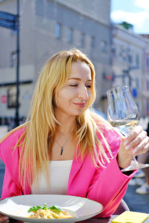 A beautiful blonde adult woman in a bright fuchsia-colored jacket sits at a table in a cafe, drinks white wine, eats spaghetti and enjoys the weather. Vertical photoの写真素材