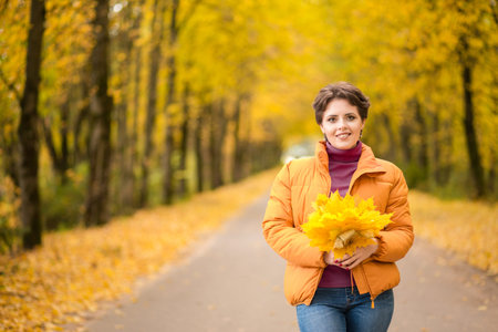 Beautiful brunette woman posing in a yellow jacket in an autumn park with a bouquet of autumn leaves. Horizontal photoの写真素材
