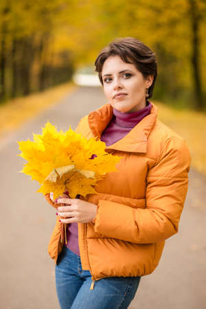 Beautiful brunette woman posing in a yellow jacket in an autumn park with a bouquet of autumn leaves and looking at the camera. Vertical photoの写真素材