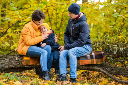 Mother, father and little son sit on a plaid on a log in the autumn park and spend time together. Horizontal photoの写真素材