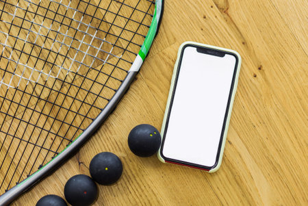 Black squash balls, a racket and a mobile phone with a putty screen lie on a parquet background. The...