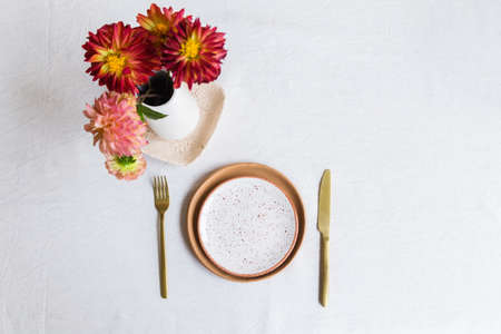A set of orange clay plates, a knife and a fork on a white tablecloth made of natural material. A vase with dahlia flowers stands nearby. Top view. Horizontal photoの写真素材