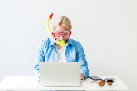 A blonde woman in a diving mask sits in the office, orders a tourist trip online, tickets for a plane, bus or train. Woman dreaming of sea at workplace. Horizontal photoの写真素材