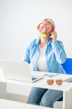 A blonde woman in a diving mask sits in the office and happily announces by phone that she is going on a trip or communicating with a tour operator. Vertical photoの写真素材