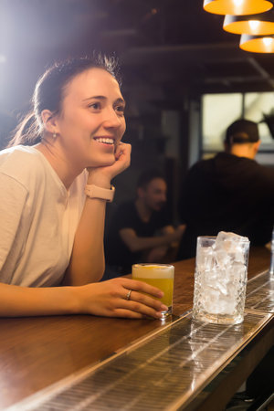 A beautiful girl is relaxing in a bar with a glass of cocktail in her hand. The girl looks at the bartender and smiles. Vertical photoの写真素材