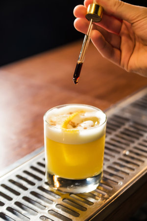 Hand of a male bartender holds a pipette and adds a drop of liquid to a glass with a cocktail on the...