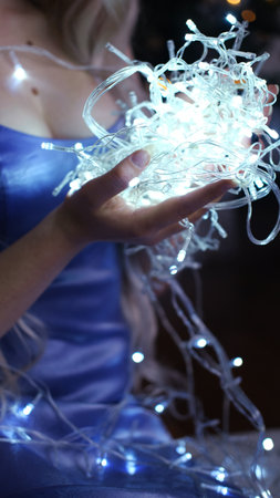 A girl in a blue dress sits on the floor and holds a luminous garland in her palms. Christmas atmosphere. Vertical photoの写真素材
