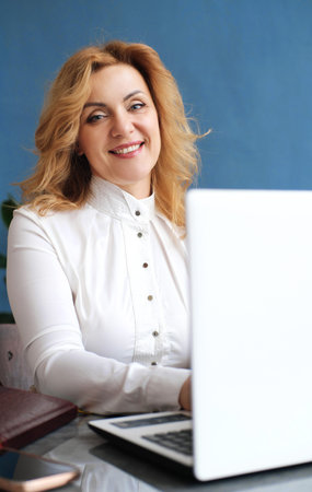 Beautiful mature blonde business woman sitting at a laptop in the office, smiling and looking at the...