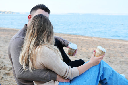 Date on the beach. A young couple in sweaters sits on a plaid, holds coffee in stacks in their hands and kisses. Horizontal photoの写真素材