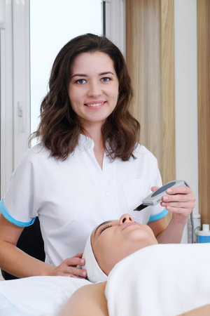 Cosmetologist aesthetist performs the procedure of ultrasonic facial cleansing. The girl looks at...