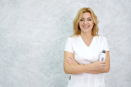 A beautiful mature woman beautician in a medical suit holds an ultrasound in her hands. Woman posing on a gray background, smiling and looking at the camera. Horizontal photoの写真素材