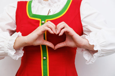 A girl in a red Russian folk dress makes a heart gesture with her fingers in front of her chest showing her love. Horizontal photoの写真素材