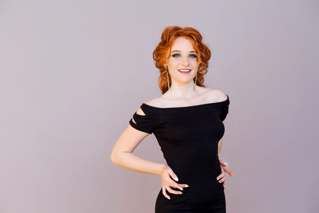 Beautiful slender red-haired girl with straight earrings in a tight dress posing on a gray background. Horizontal photoの写真素材