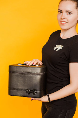 A brunette girl in a black T-shirt demonstrates a black case for beauty masters on a yellow...