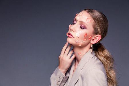 Beautiful woman in a jacket posing with flower petals on her face on a gray background. Femininity...