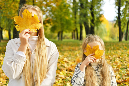 The family spends time together in the park. Girl and mother sit on a blanket and cover their faces with autumn leaves. Horizontal photoの写真素材