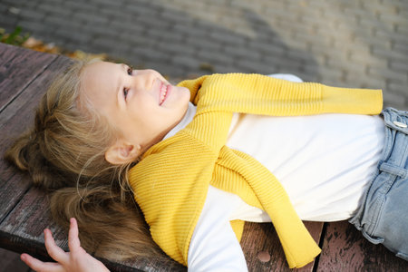 A little blonde preschool girl in jeans, a turtleneck and a sweater lies on a bench and looks up. Horizontal photoの写真素材