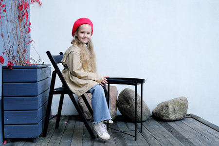 A beautiful little girl in a trench coat, red beret and jeans poses while sitting at a table in a street cafe. Girl sitting with her back to the camera. Horizontal photoの写真素材