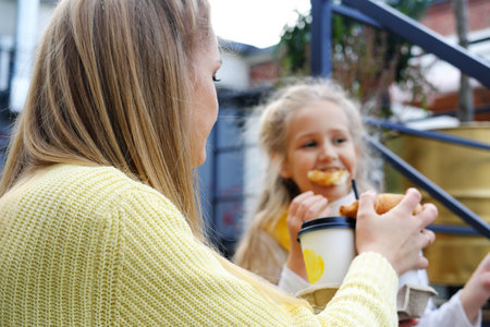 Mom and daughter are sitting on the steps with glasses of hot chocolate and croissants. Mother feeding her daughter with a croissant. Horizontal photoの写真素材