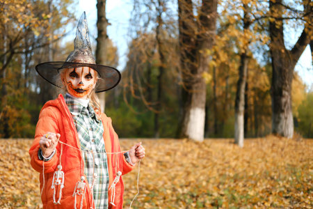 A child girl in an orange jacket, a witch's hat and with orange makeup on her face holds a...